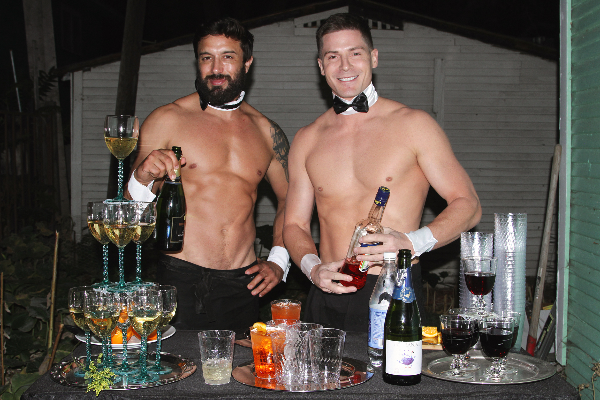 Two shirtless white male butlers smiling behind a bar